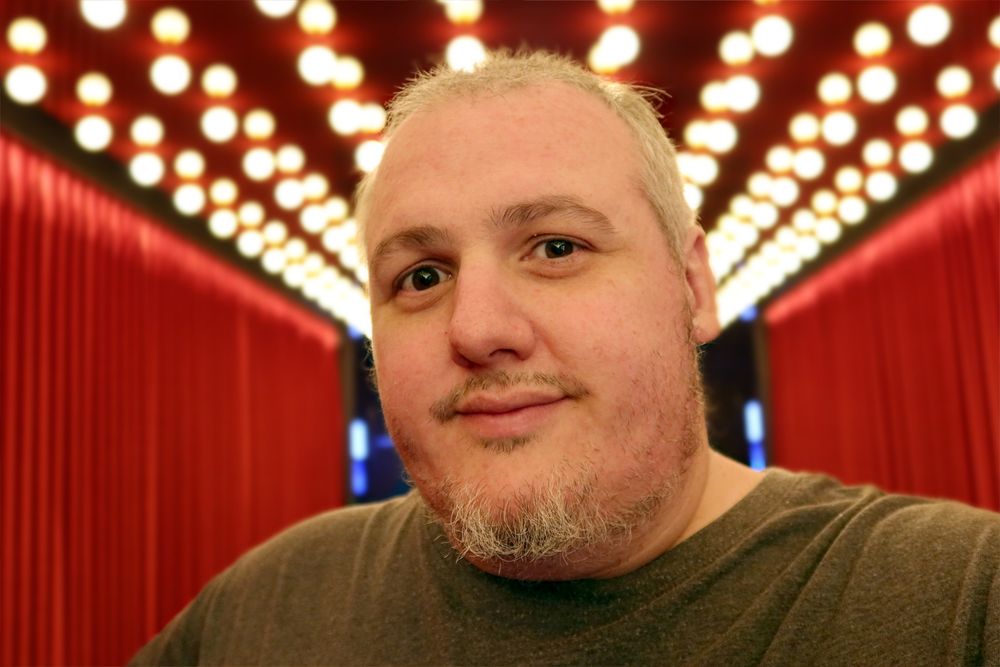 Spammals face smiles to the camera. Behind him are red curtails to the left and right. The ceiling has lots of glowing light bulbs