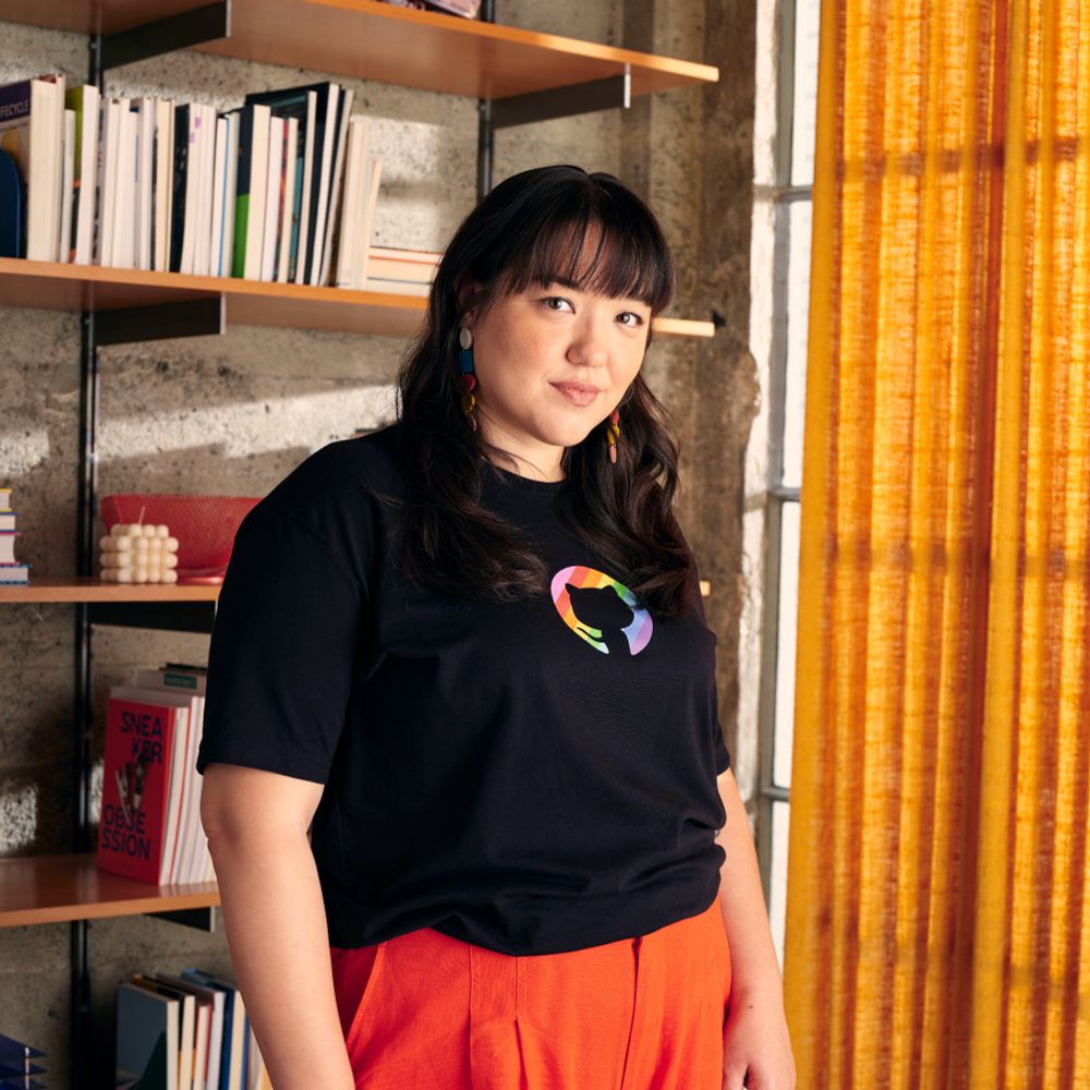 Person standing in front of a bookshelf, wearing a black t-shirt with the GitHub logo with Pride colors, and bright orange pants, smiling slightly at the camera.