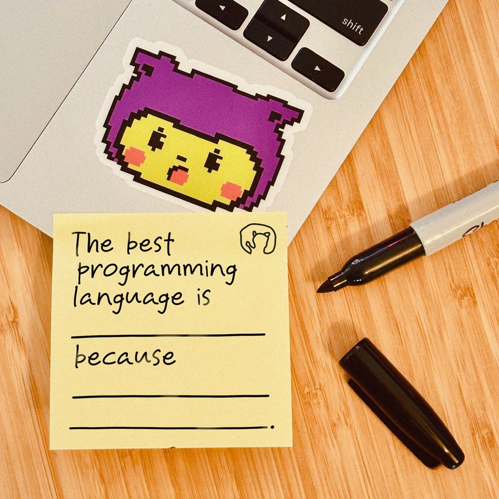 A sticky note on a laptop that reads "The best programming language is _ because _". There is an octocat sticker on the laptop. An open pen is laying on the table beside the laptop. 