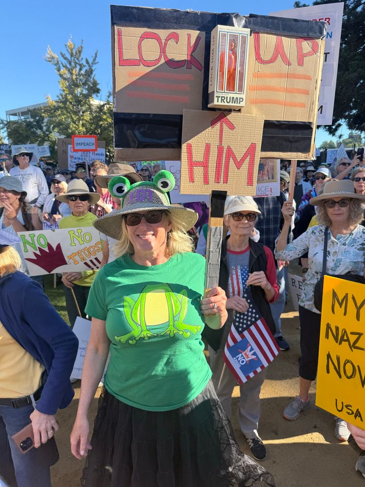 Woman in frog shirt holding a “Lock Him Up” protest sign. 