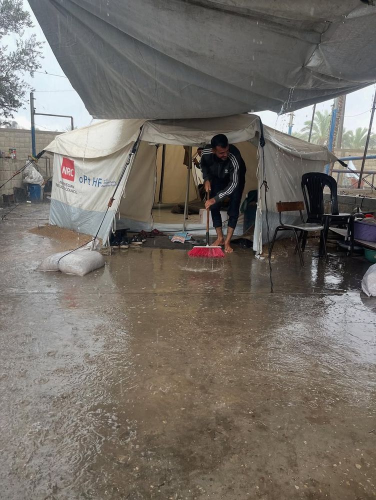 Ahmed sweeping the standing water away from the entrance of his tent, so it does not flood the interior any worse