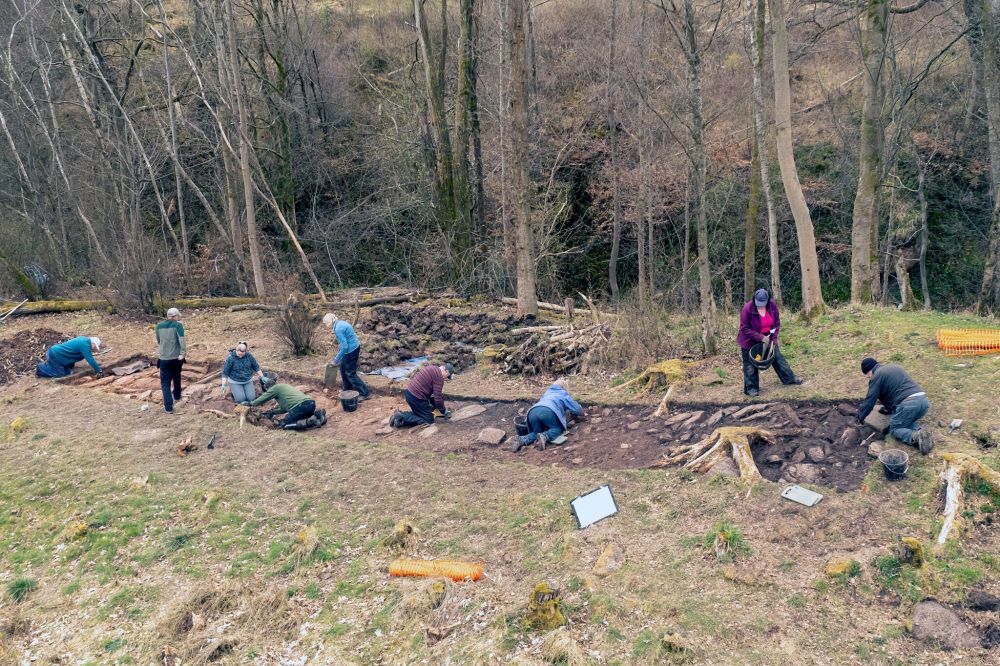 A big group of people wearing outdoor clothes spread out working in an archaeological trench in the forest. Clutha’s dig at Avon Gorge, run by Archaeology Scotland April 2023. © E. Smith