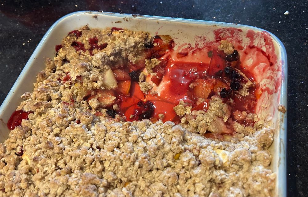 A plum and apple crumble dessert in a ceramic dish. A portion of the crumble has been served revealing a deep red fruit liquor at the bottom of the dish