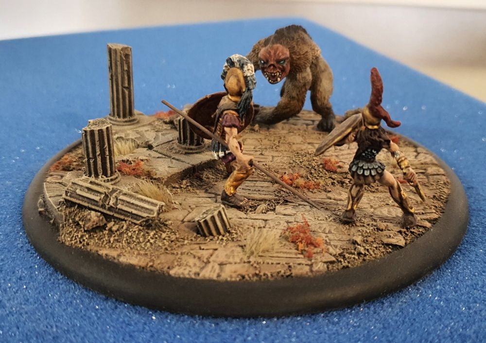Model diorama of two Greek adventurers facing a Pumpkin-headed bear creature in amongst ruins.