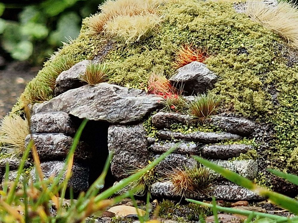 Photo of a finished barrow mound model, seen through a foreground of clumped grass. A dark stone doorway leads into a tussocked hillside...