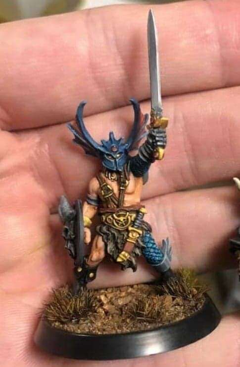 Model of a barbarian in hero harness with a winged helm and one arm and one leg armoured in blue scale. His sword is raised to the asky in a challenge.