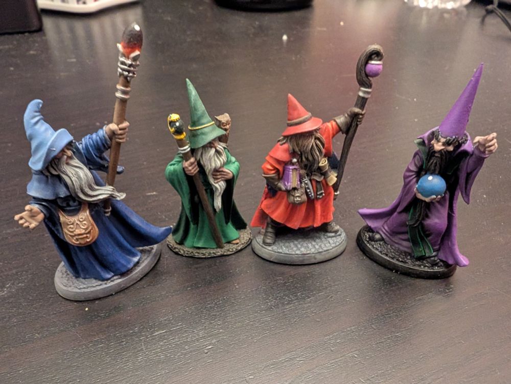 Four miniature painted wizards with pointy hats. The leftmost one is blue, then (moving right) green, red/orange, and purple.