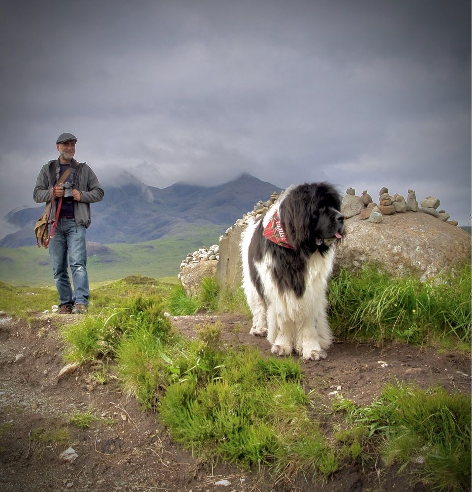Wolkenverhangener Himmel, im Hindergrund Berge, grünes Gras. Der schwarz-weiße Neufundländer steht vor einem Fels auf dem kleine Steintürmchen zu sehen sind. Link im Bild ein paar Schritte weiter hinten stehe ich und halte eine Kamera in der Hand.