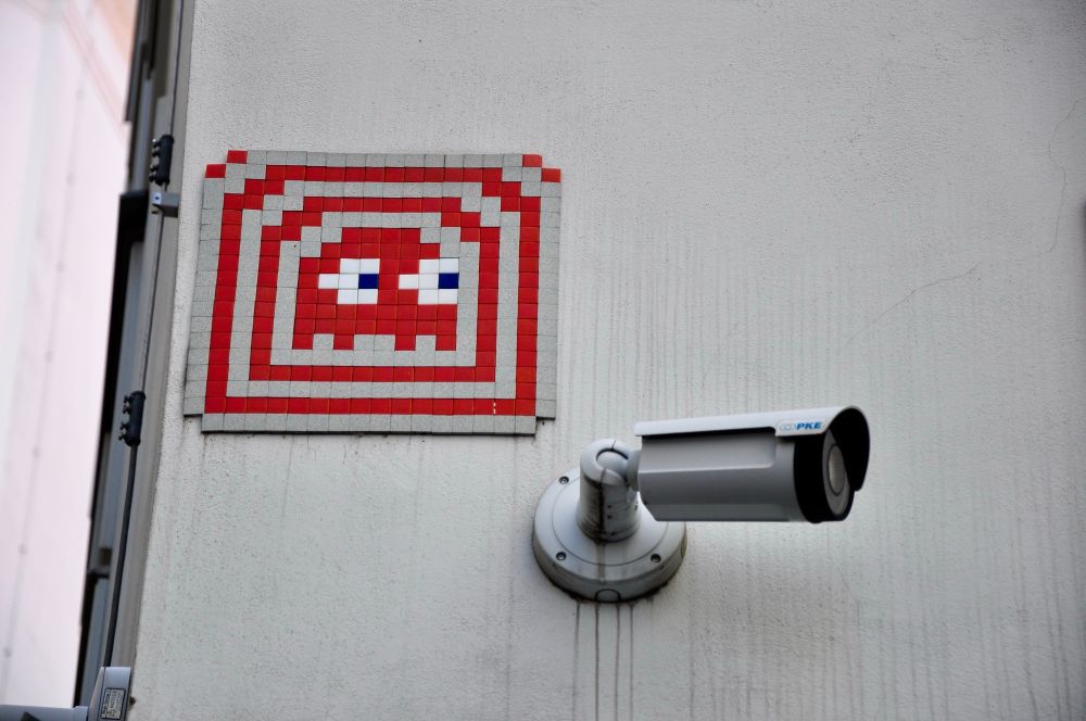 The color image shows a wall with a small surveillance camera mounted on it and a pixel art mosaic above. The mosaic resembles a space invader from the classic video game, created with red, white, blue, and gray tiles. The extraterrestrial appears to be looking sideways, seemingly the same direction as the security camera below.