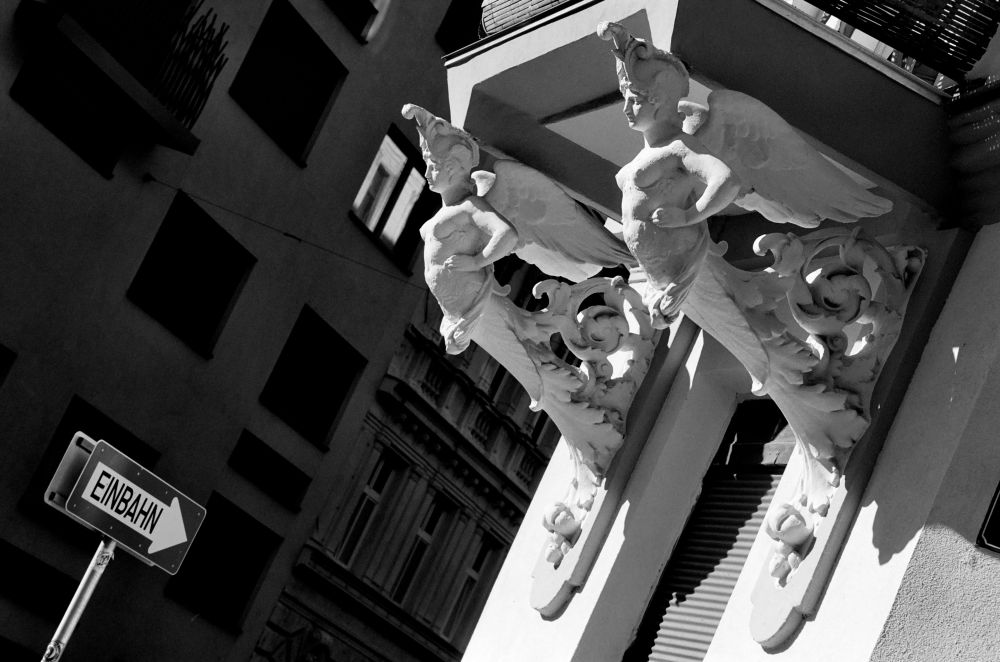 The black and white image shows two ornate sculpted figures with wings and elaborate helmets supporting the corner of a building. Their bodies merge into decorative scrollwork. In the background, a tilted “Einbahn” (one-way) street sign with an arrow points to the right against the facade of another building.