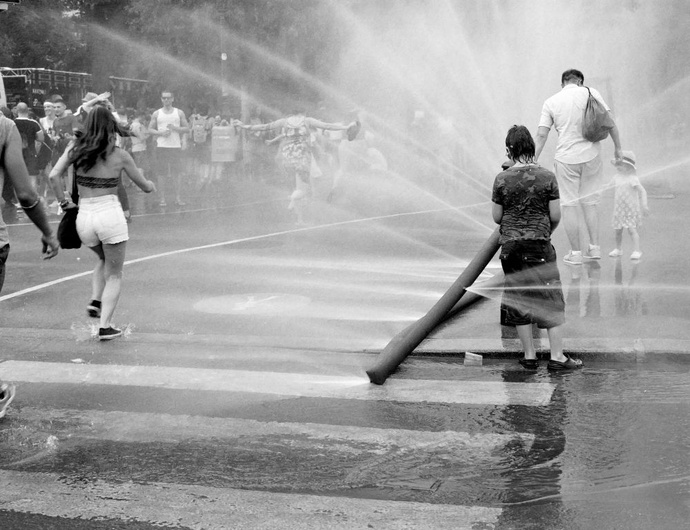The black and white image captures a lively street scene where people, including children and adults, are playing and walking through water sprayed from a large hose. Water jets crisscross the air, creating a misty atmosphere filled with movement, splashes, and laughter. A boy stands near the hose, while others move joyfully through the cool spray on a hot day.