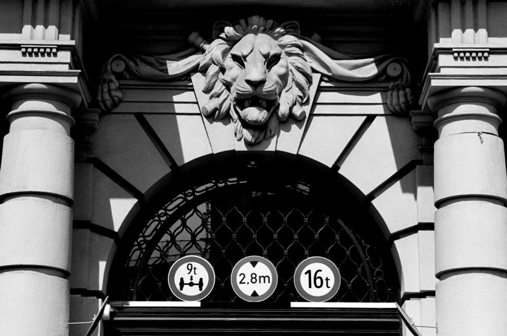 The black and white image shows an ornate architectural entrance featuring a carved lion’s head above a large arched doorway. Below the arch are three circular traffic signs indicating a 9-ton axle load limit, a 2.8-meter height restriction, and a 16-ton total weight limit.