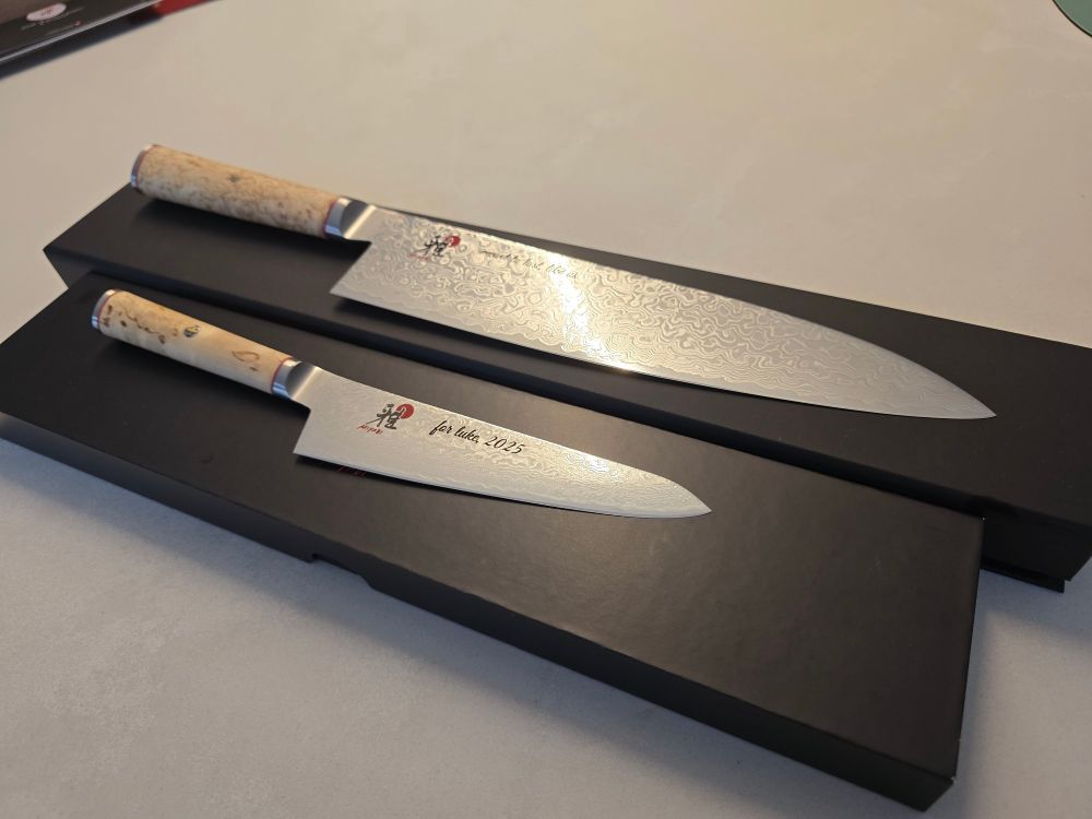2 Miyabi Japanese kitchen knives, one Gyotoh and one Shotoh, on the boxes they came in. Both have Damascus patterns in their blades and Karelian Birch wood handles with bold swirling grain.