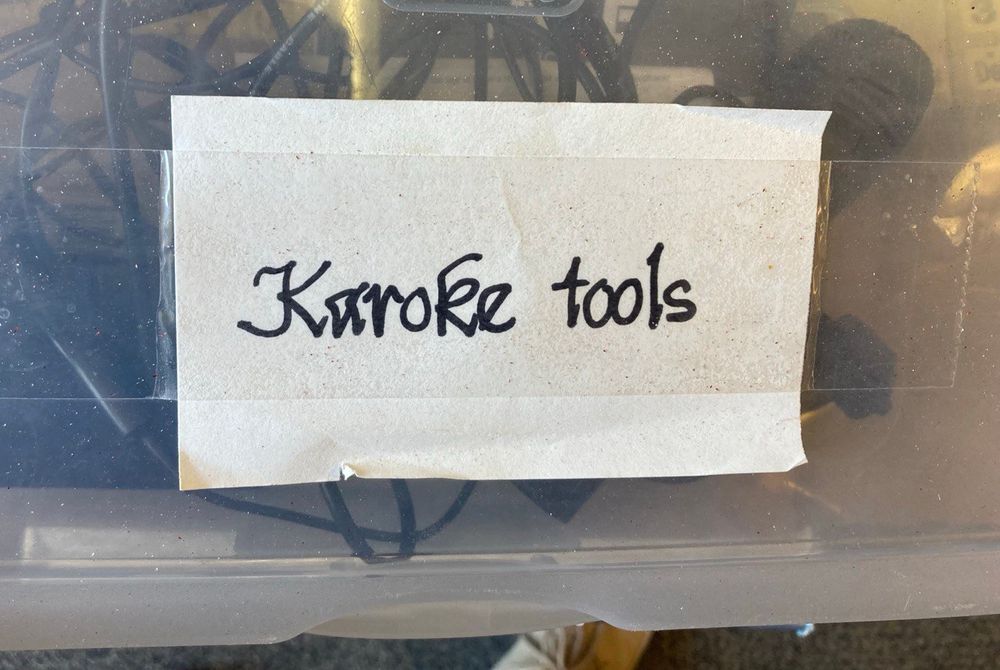 A photograph of a plastic storage box with various black cords and cables inside. A paper with "Karoke tools" (sp) in a handwritten Chancery font is taped to it.