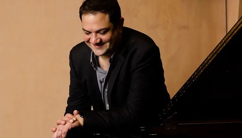 An image of a man, Coady Green, with short black hair in a black suit and grey collared shirt with an open collar. He leaning over a piano, looking down and smiling.