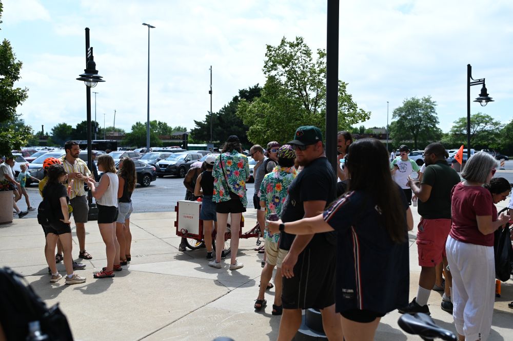 A large number of people milling around the sidewalk by the ice cream cart