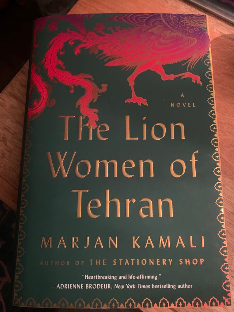 Photo of the novel “The Lion Women of Tehran” on the edge of a desk. The cover/jacket of the novel has a purple/pink/orange phoenix at the top above the gold lettering of the title and author name (Marjan Kamali: Author of the stationary shop) all on a green background with gold border 