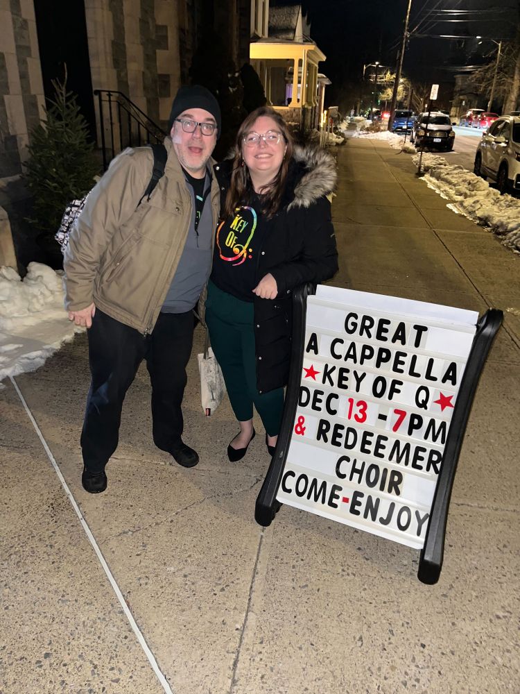 A couple of posers next to a sandwich board which says
GREAT
A CAPPELLA
* KEY OF Q *
DEC 13 - 7PM
& REDEEMER
CHOIR
COME-ENJOY
