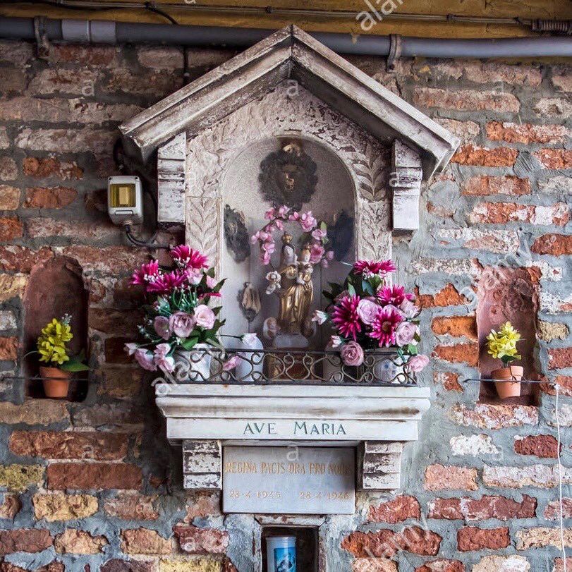 A decorative wall shrine featuring a statue of a saint in an alcove, with the inscription "AVE MARIA" and a memorial plaque below it.