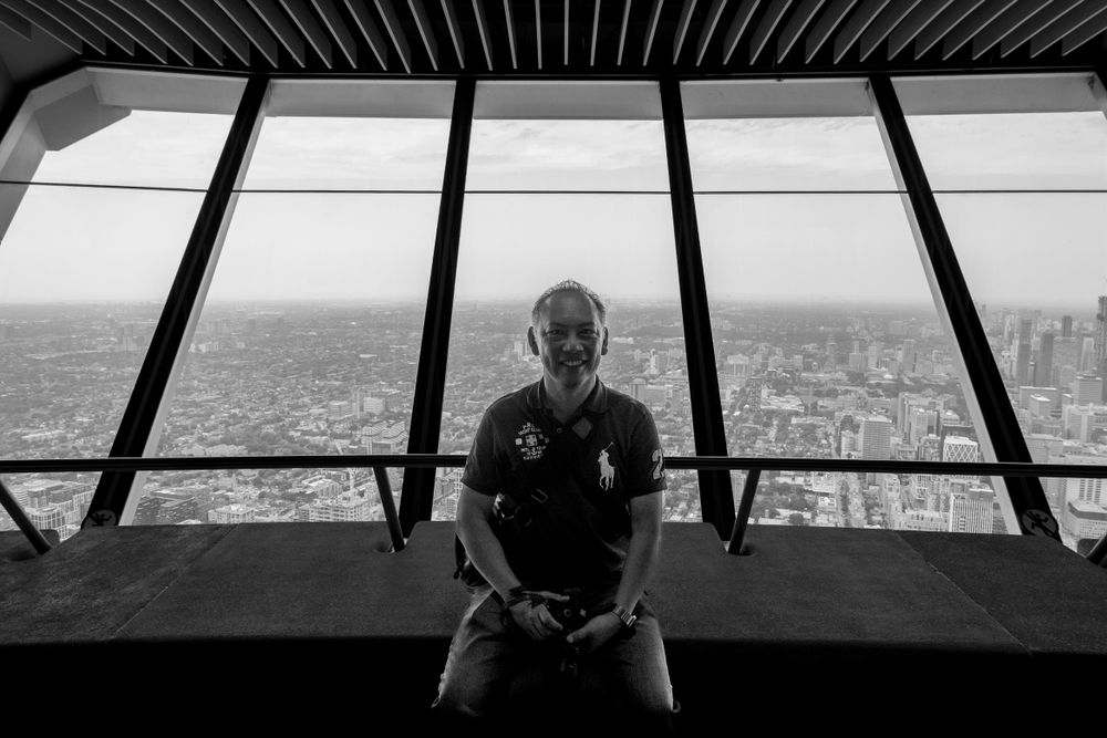 A photo of me (Asian man) sitting at the window of Toronto’s CN Tower. 