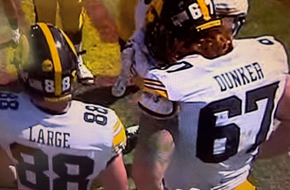 Two Iowa Hawkeye football players in the offensive huddle, facing away from the camera so we can see the names LARGE and DUNKER on the backs of their jerseys