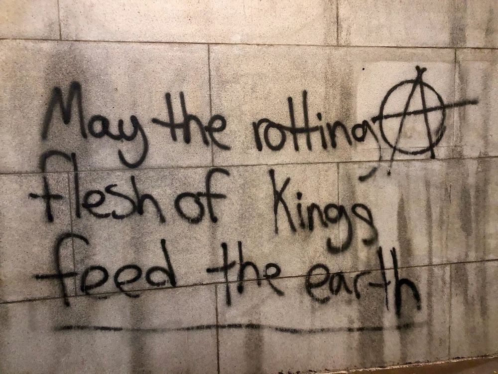 The words MAY THE ROTTING FLESH OF KINGS FEED THE EARTH with an anarchy symbol, spray painted on a concrete wall.