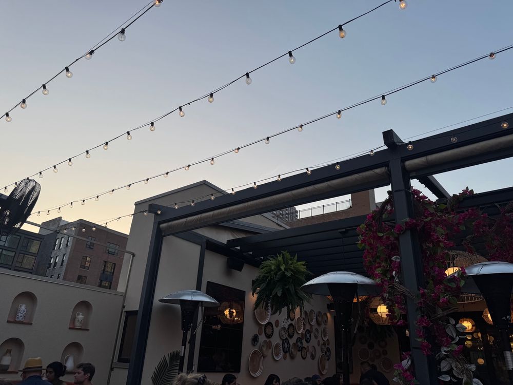 City scene at dusk. Strings of light above a tan stucco wall. The wall has ceramics in alcoves and wicker, embroidered in serving pieces. There are flowers hanging from a pergola. Buildings in the background. 