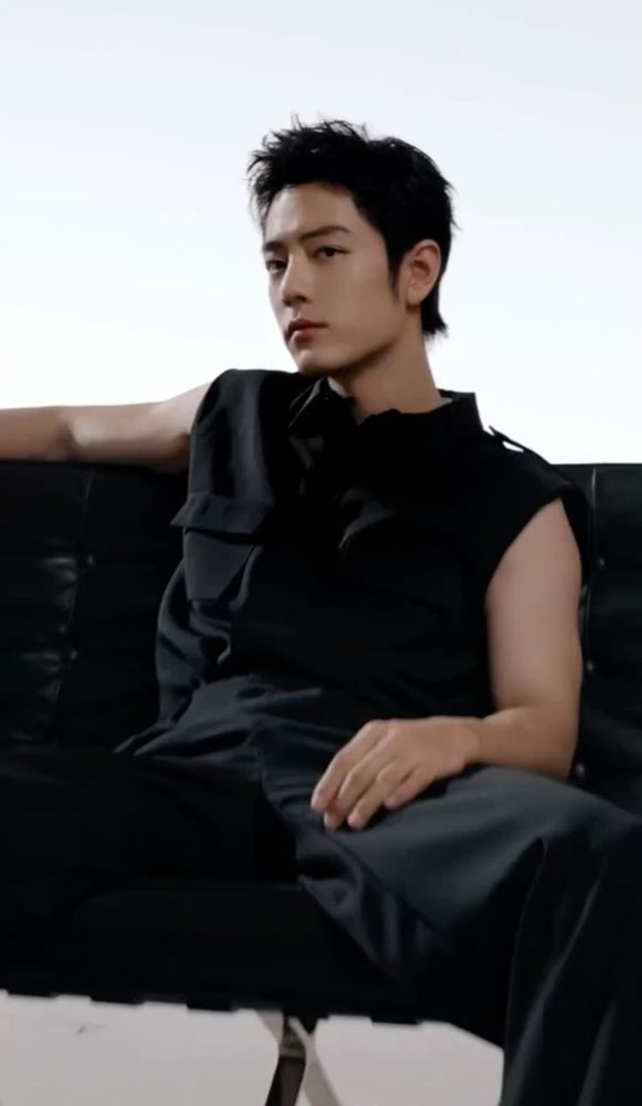 Xiao Zhan gazing into the camera in black pants and a sleeveless black shirt sitting on a black couch with his legs spread, his left hand over his tight, the other over the back of the couch. 