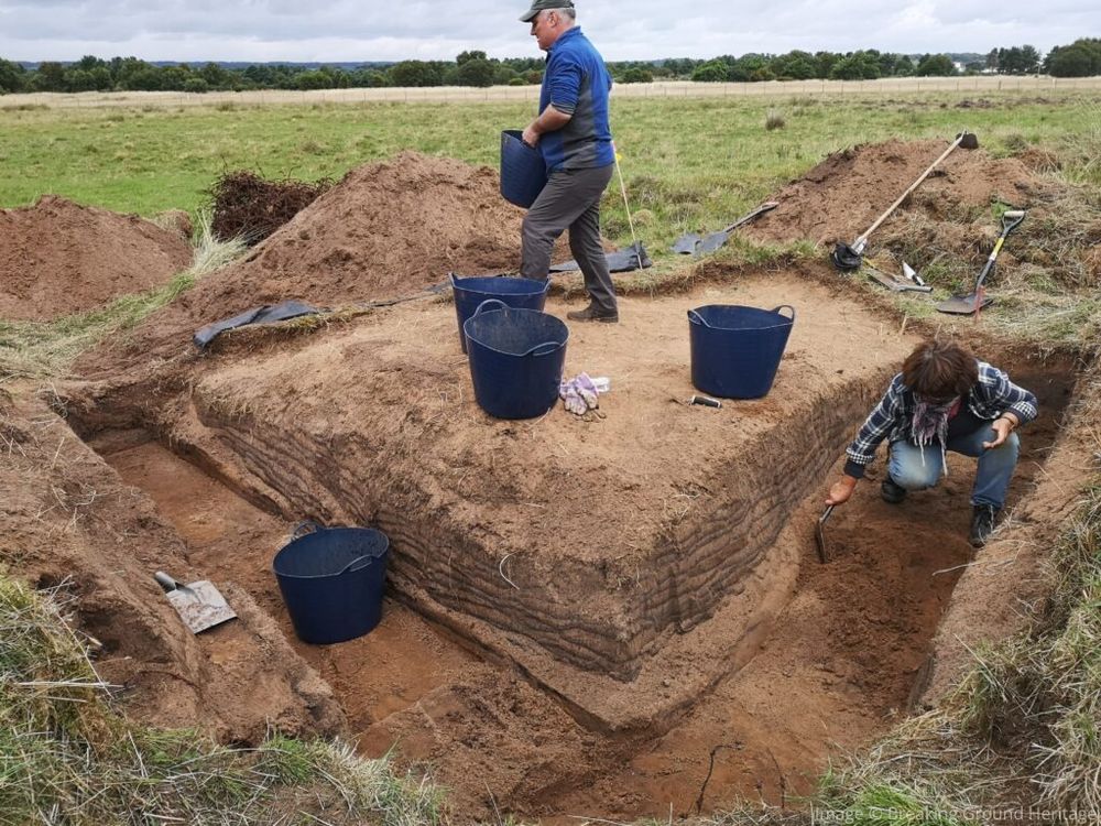 An L-shaped trench with one person on top holding a bucket and one person in the trench digging 