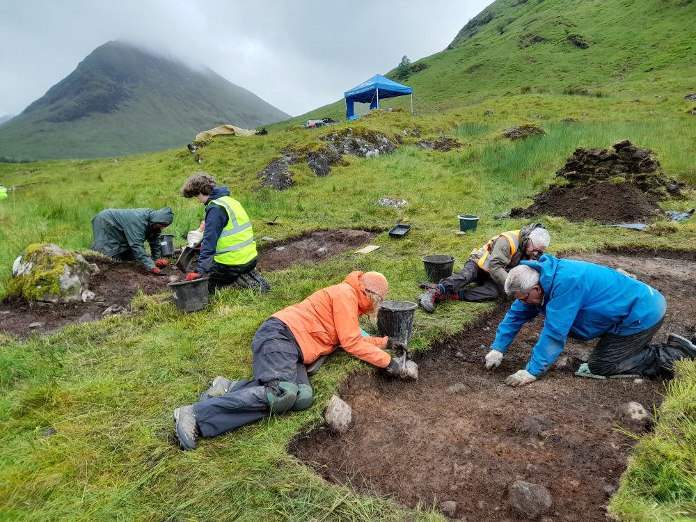 Several people in trenches in a mountainous setting with buckets and piles of dirt near each trench  