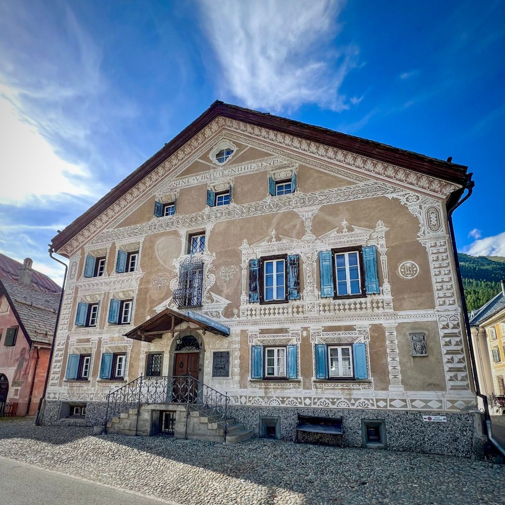 Ein grosses Engadiner Haus mit reich verzierter Fassade steht an einer gepflasterten Strasse. Die Fassade ist mit weissen Sgraffiti auf hellbraunem Putz geschmückt und zeigt Ornamente, Fensterrahmungen und einzelne bildhafte Darstellungen. Die Fensterläden sind blau, die Eingangstür liegt erhöht und ist über eine steinerne Treppe mit schwarzem schmiedeeisernem Geländer erreichbar. Über dem mittleren Fenster im Giebeldreieck steht ein lateinischer Schriftzug. Der Himmel ist blau mit vereinzelten Wolken, links und rechts sind angrenzende Häuser sichtbar.