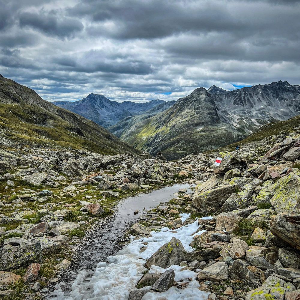 Blick vom Scalettapass über eine steinige, nasse Berglandschaft mit Resten von Altschnee im Vordergrund. Ein schmaler Wanderweg mit Pfützen führt durch Geröll und grüne Hänge in ein weites Tal. Im Hintergrund ragen graue und grüne Bergketten unter einem dramatisch bewölkten Himmel auf. Auf einem Fels ist eine rot-weisse Wanderwegmarkierung aufgemalt.