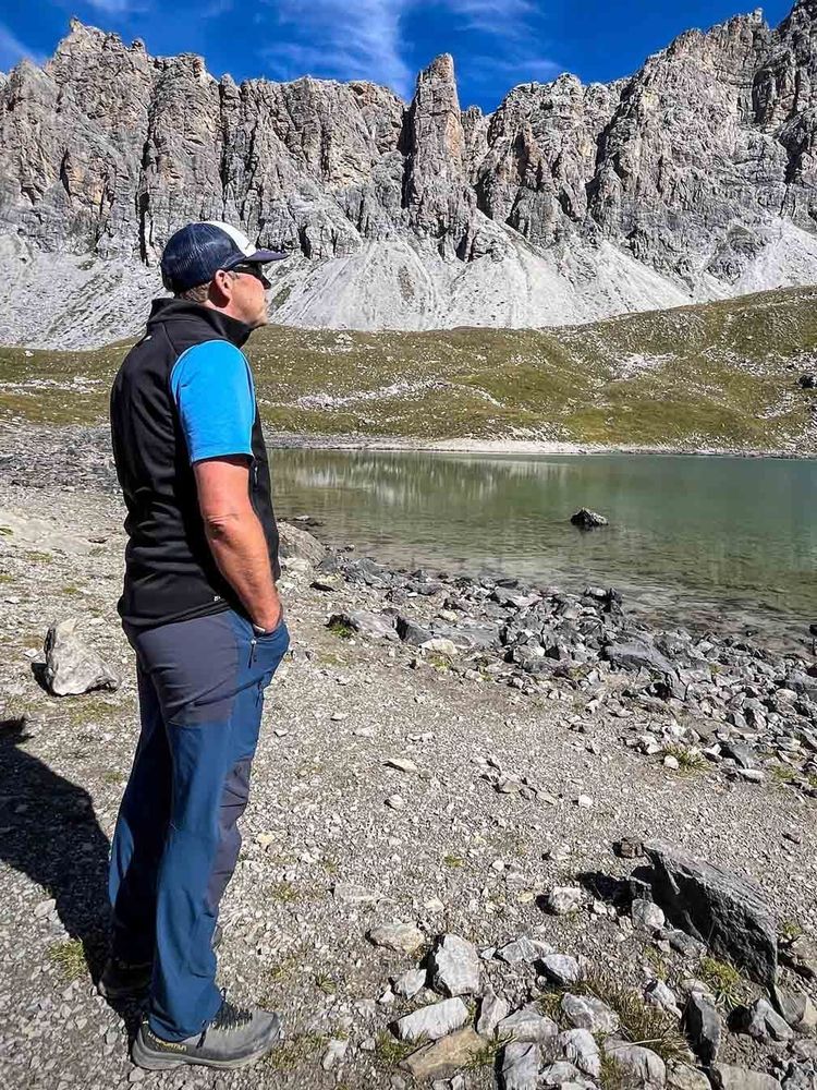 Ein Mann steht seitlich am Ufer eines Bergsees und blickt zu den steilen, grauen Felszacken auf der gegenüberliegenden Seite. Er trägt Wanderhose, Wanderschuhe, blaues Shirt, schwarze Weste und Cap. Der Himmel ist blau mit leichten Wolken, der Boden ist steinig.