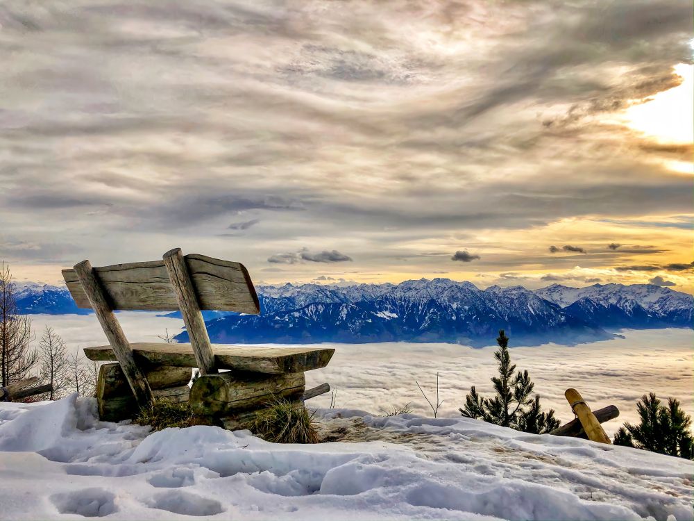 Eine verwitterte Holzbank steht auf einer kleinen, ebenen Schneefläche oberhalb eines dichten Nebelmeers. Der Schnee rund um die Bank ist sichtbar zertrampelt. Im Hintergrund zieht sich eine lange Kette hoher, verschneiter Alpenzüge über den Horizont. Darüber liegt ein dramatischer Himmel mit dichten Wolken und warmen Lichtreflexen am rechten Bildrand. Rechts im Vordergrund stehen ein kleiner Baum und einige verschneite Äste.