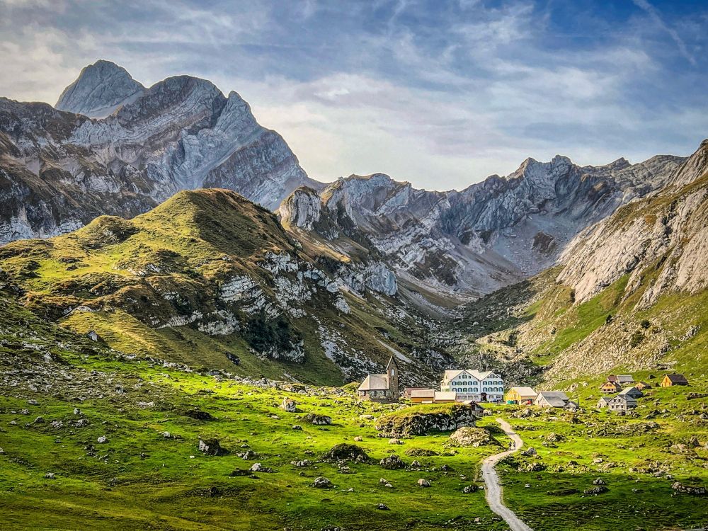 Blick auf die Meglisalp im Alpstein: Im Vordergrund eine grüne Alpweide mit verstreuten Steinen und einem Weg, der zu den hellen Häusern und Hütten der Siedlung führt. Dahinter ragen steile, graue Felswände und Berggipfel auf, die von der weichen Herbst-Morgensonne beleuchtet werden. Der Himmel ist leicht bewölkt und verstärkt das warme Licht auf den Berghängen.