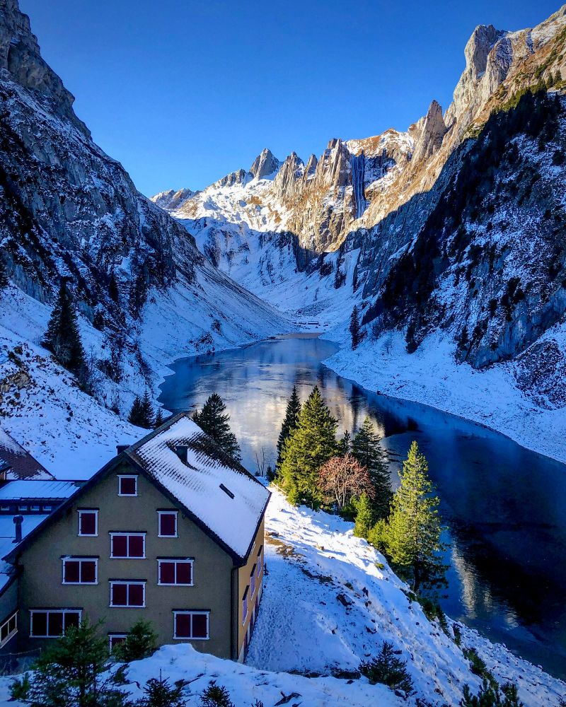 Ein winterliches Bergpanorama am Fälensee im Alpstein. Im Vordergrund steht das Berghaus Bollenwees mit schneebedecktem Dach. Dahinter liegt der teilweise zugefrorene Fälensee, umrahmt von steilen, schattigen Felswänden. Im Hintergrund leuchten die schneebedeckten Gipfel in der Sonne, während einzelne Tannen am Seeufer im Licht hervorstechen.