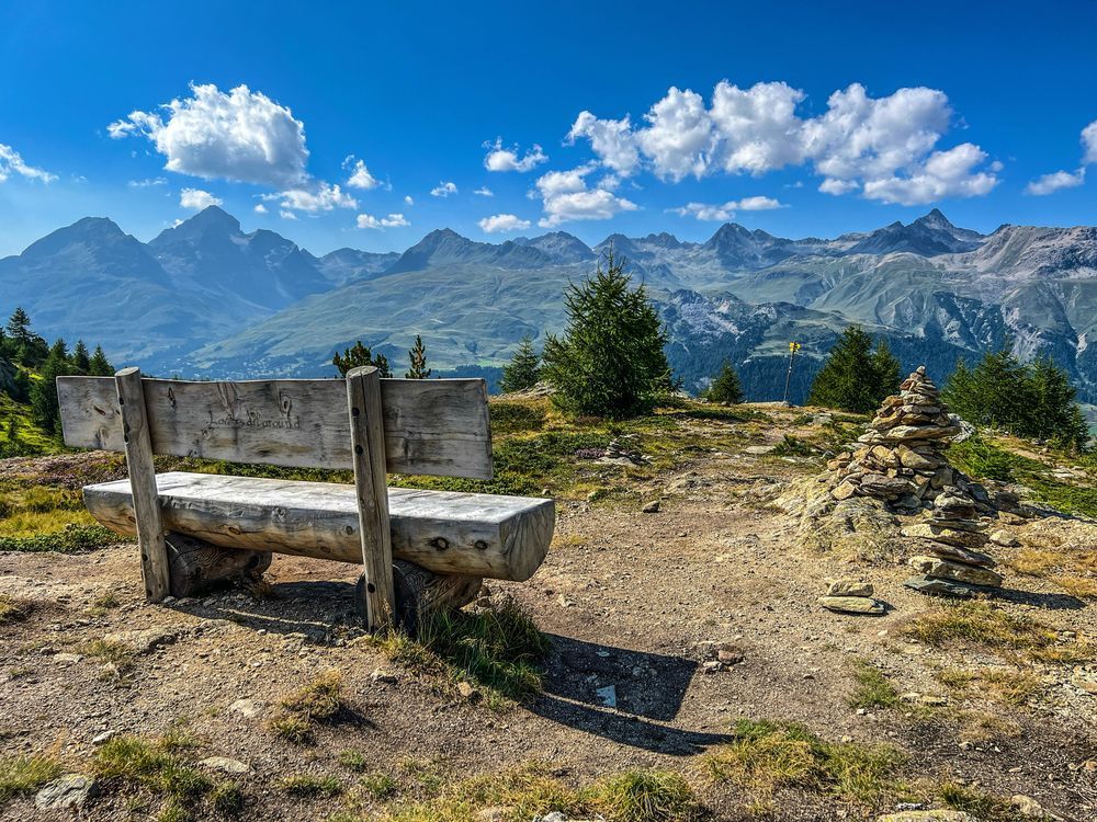 Holzbank auf einer sonnigen Anhöhe mit Blick auf die Engadiner Alpen. Links im Bild steht die rustikale Bank, auf deren Lehne «Love is all around» eingeritzt ist. Rechts daneben ein kleiner Steinmann aus aufgeschichteten Steinen. Im Hintergrund grüne Hänge, Berggipfel und ein blauer Himmel mit einzelnen weissen Wolken.