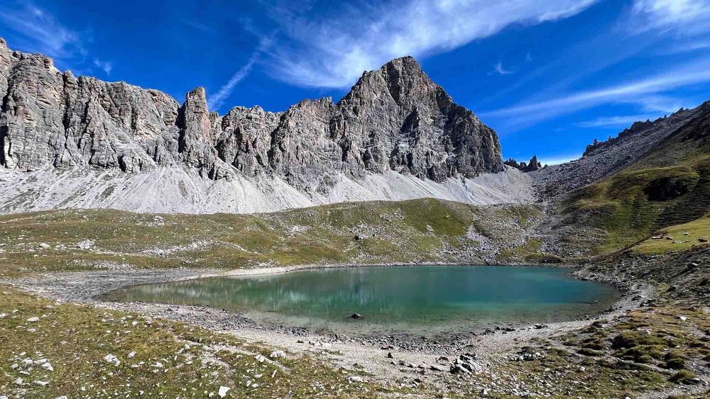 Ein türkisgrüner Bergsee liegt in einer offenen, steinigen Hochebene unterhalb einer imposanten, grau-weissen Felswand mit schroffen Zacken. Der Himmel ist klar blau mit einzelnen Wolkenstreifen. Links und rechts steigen Geröllhänge an.