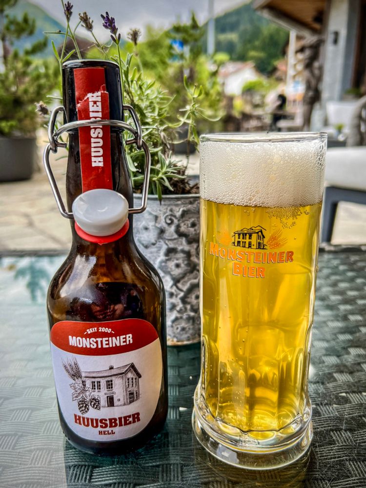 Flasche und Glas Huusbier aus Monstein auf sonniger Terrasse in Klosters, im Hintergrund Pflanzen und ein traditionelles Haus.