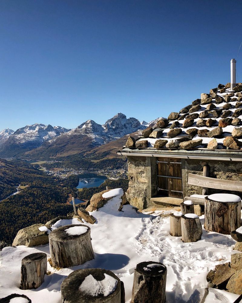 Blick von der schneebedeckten Terrasse der Segantinihütte auf 2731 Metern ins Oberengadin. Im Vordergrund stehen runde Holzstümpfe als Sitzgelegenheiten, rechts ist die steinerne Aussenwand der Hütte mit einer Holztür zu sehen. Im Hintergrund das herbstlich gefärbte Tal mit dem St. Moritzersee und dem Stazersee sowie den Gipfeln von Piz Albana und Piz Julier unter wolkenlosem Himmel.