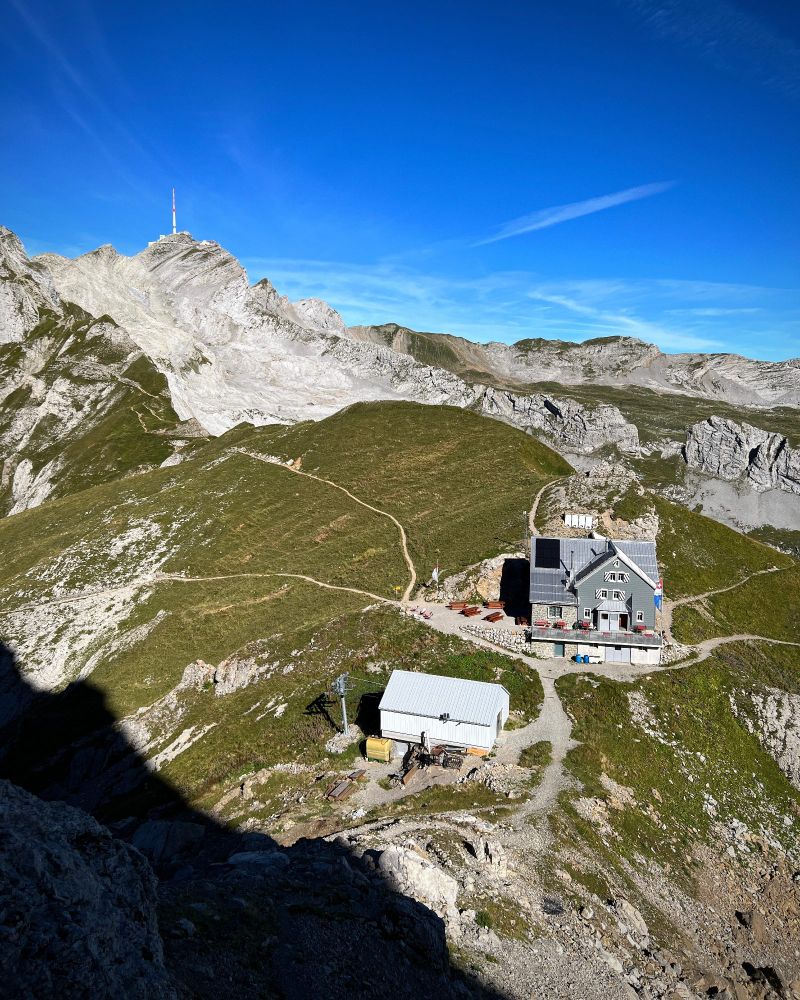 Blick von oben auf das Berggasthaus Rotsteinpass im Alpstein, umgeben von grünen Wiesen und grauen Felsflanken. Links im Hintergrund ragt der markante Säntisgipfel mit seiner Antenne in den tiefblauen Himmel. Ein schmaler Wanderweg schlängelt sich vom Pass über die Hänge, im Vordergrund liegt der Schatten einer steilen Felswand.