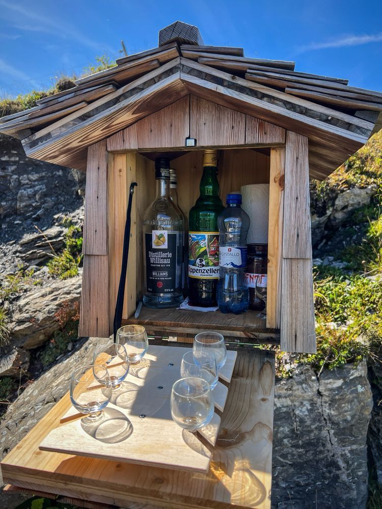 Ein hölzernes Kästchen in Form eines kleinen Häuschens mit Schindeldach steht im Gebirge. Darin sind mehrere Spirituosenflaschen, eine Wasserflasche und Küchenpapier verstaut. Vor dem Kasten ist ein Brett mit sechs kleinen runden Trinkgläsern angebracht. Im Hintergrund sind Felsen, Gras und blauer Himmel zu sehen.