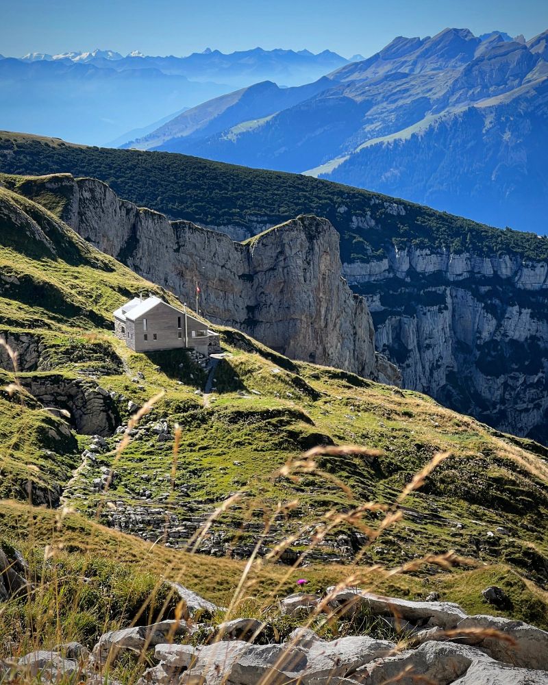 Blick vom Zwinglipass auf die Zwinglipasshütte mit Schweizer Flagge, die einsam auf einer grasbewachsenen Hochterrasse steht. Im Hintergrund ragen steile Kalkwände empor, dahinter erstrecken sich mehrere Gebirgsketten mit markanten Gipfeln. Im Vordergrund unscharfe Gräser, die im Sonnenlicht golden leuchten. 