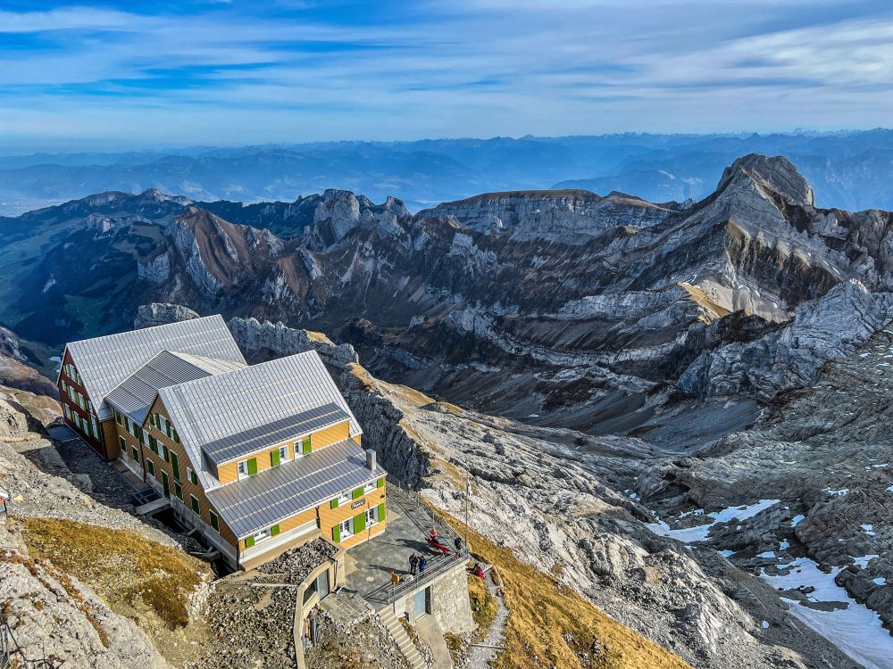 Auf dem Foto ist das Berghotel Alter Säntis auf dem Säntisgipfel zu sehen. Das grosse, mehrteilige Gebäude mit grün-weissen Fensterläden steht auf felsigem Untergrund und fällt durch seine gelbbraune Fassade und das silbergraue Dach auf. Im Hintergrund breitet sich die markante Felslandschaft des Alpsteins aus, mit tiefen Tälern, schroffen Felswänden und Gipfeln wie dem markanten Altmann. Der Himmel ist klarblau, die Fernsicht reicht bis zu den Alpen am Horizont.