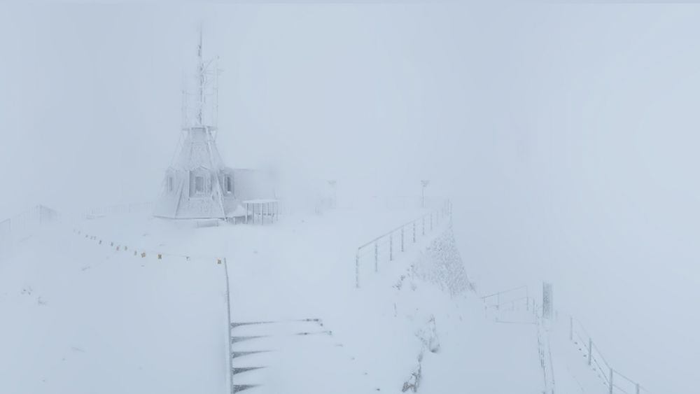 Screenshot der Webcam der Säntisbahn. Zu sehen ist ein schneebedeckter Aussichtspunkt auf dem Säntis. Eine Plattform mit Geländer führt zu einer kleinen, vereisten Turmstruktur. Dichter Nebel umhüllt die Szene, sodass die Umgebung nur schemenhaft sichtbar ist. Alles erscheint in Grau und Weiss.