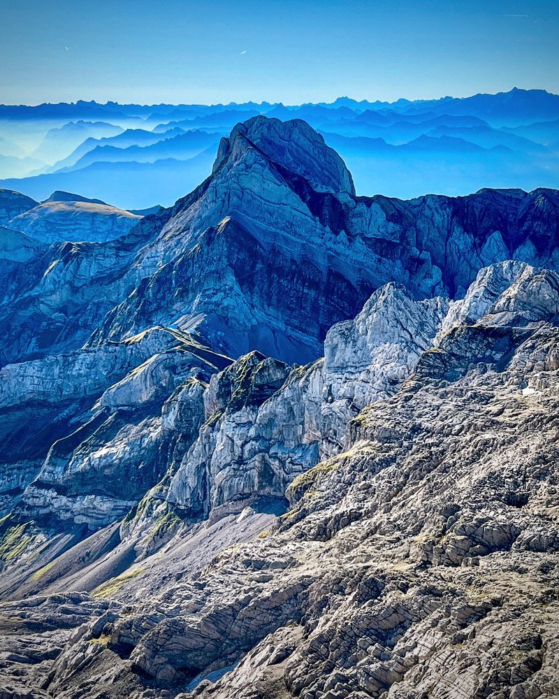 Blick auf den markanten Altmann im Alpstein, aufgenommen bei klarer Morgenstimmung. Im Vordergrund schroffe Felsformationen, dahinter der Altmann mit steilen Flanken. Am Horizont ziehen sich bläulich gefärbte Bergketten bis in die Ferne, darüber ein wolkenloser Himmel.