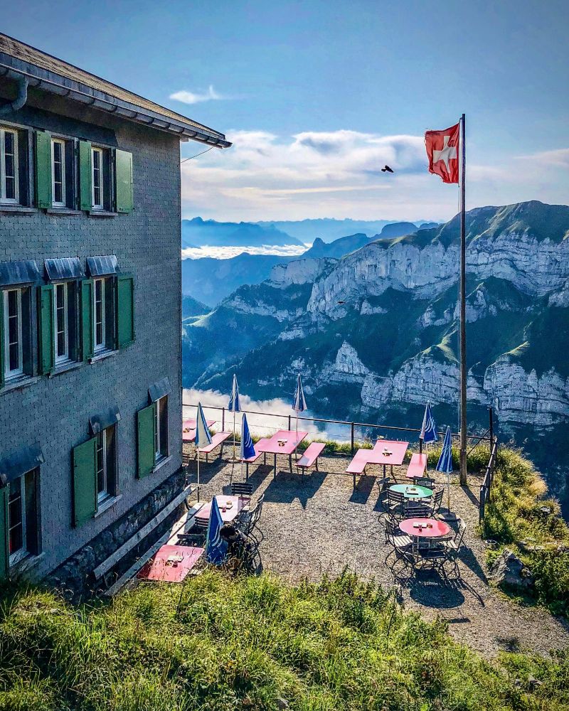 Terrasse des Berggasthauses Schäfler im Alpstein mit roten Tischen und geschlossenen Sonnenschirmen. Rechts weht eine Schweizer Fahne im Wind. Im Hintergrund fallen steile, grün-graue Felswände ab, dahinter liegt ein Nebelmeer im Tal. Eine Bergdohle fliegt über die Szene, die in kühles Morgenlicht getaucht ist. Links im Bild ist die Hausfassade mit grünen Fensterläden zu sehen.