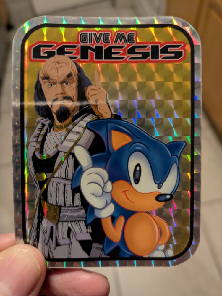 A holographic sticker featuring sonic the hedgehog and Kruge from the film Star Trek 3. The top of the sticker says, "give me genesis" in the Sega fonts. This is a line Kruge said in the movie