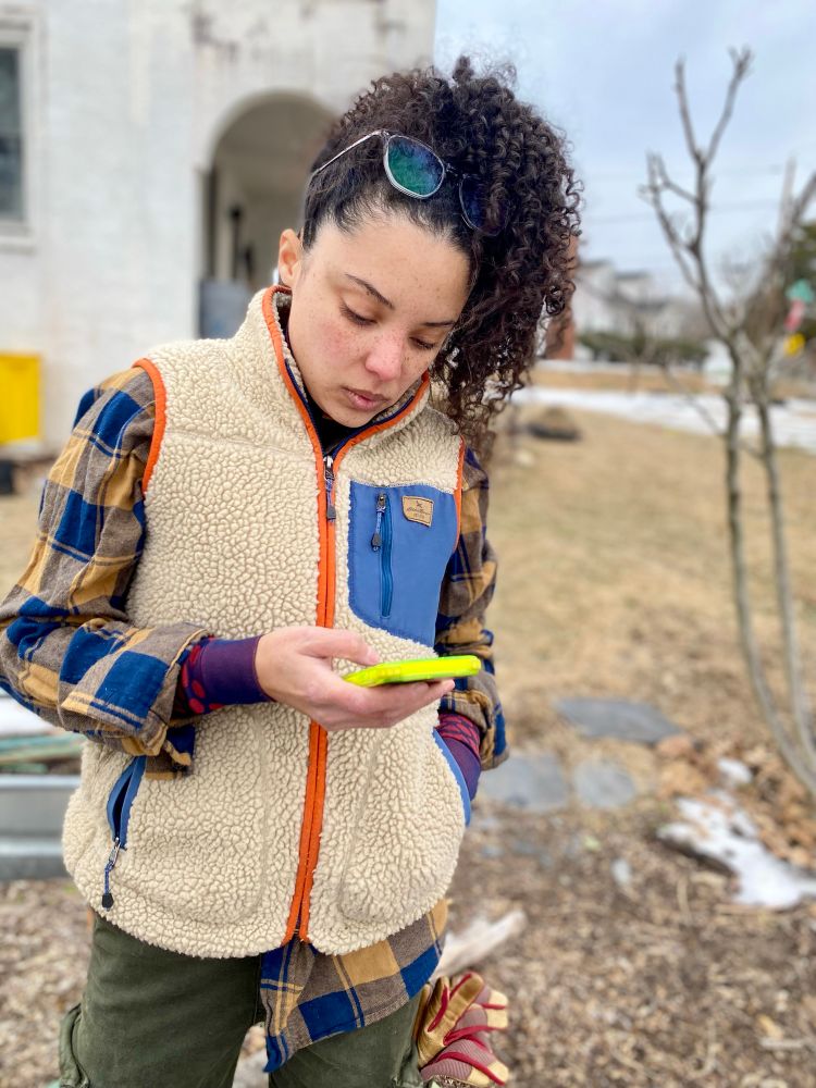 Curly haired freckle-faced woman with glasses on top of her head looking down at her phone.  Plaid shirt and fleece vest, work gloves sticking out if a pocket. A Spanish revival house in the background.