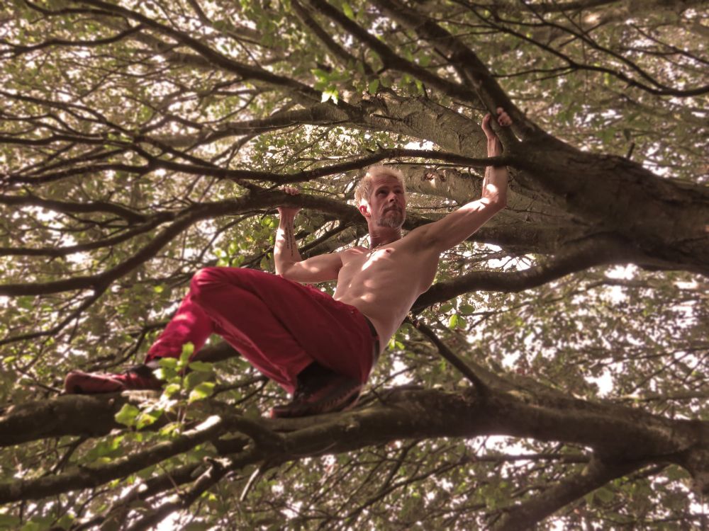 Max resting their feet on a branch while leaning backwards and holding two other branches. They are wearing red trousers and no shirt. They look pretty ripped if you ask me